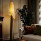 A dark-bodied Clérachic floor lamp stands lit beside a large potted palm against a panelled wall.