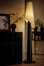 A black-bodied Clérachic floor lamp stands lit beside a dark cabinet holding a small dried-flower arrangement.