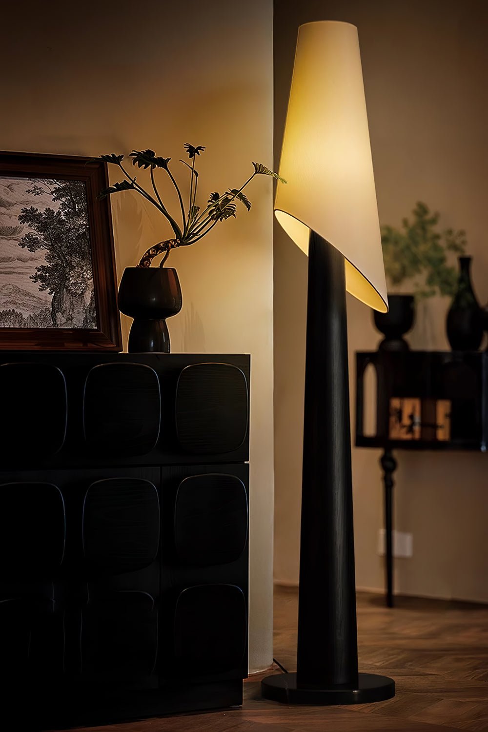 A black-bodied Clérachic floor lamp stands lit beside a dark cabinet holding a small dried-flower arrangement.