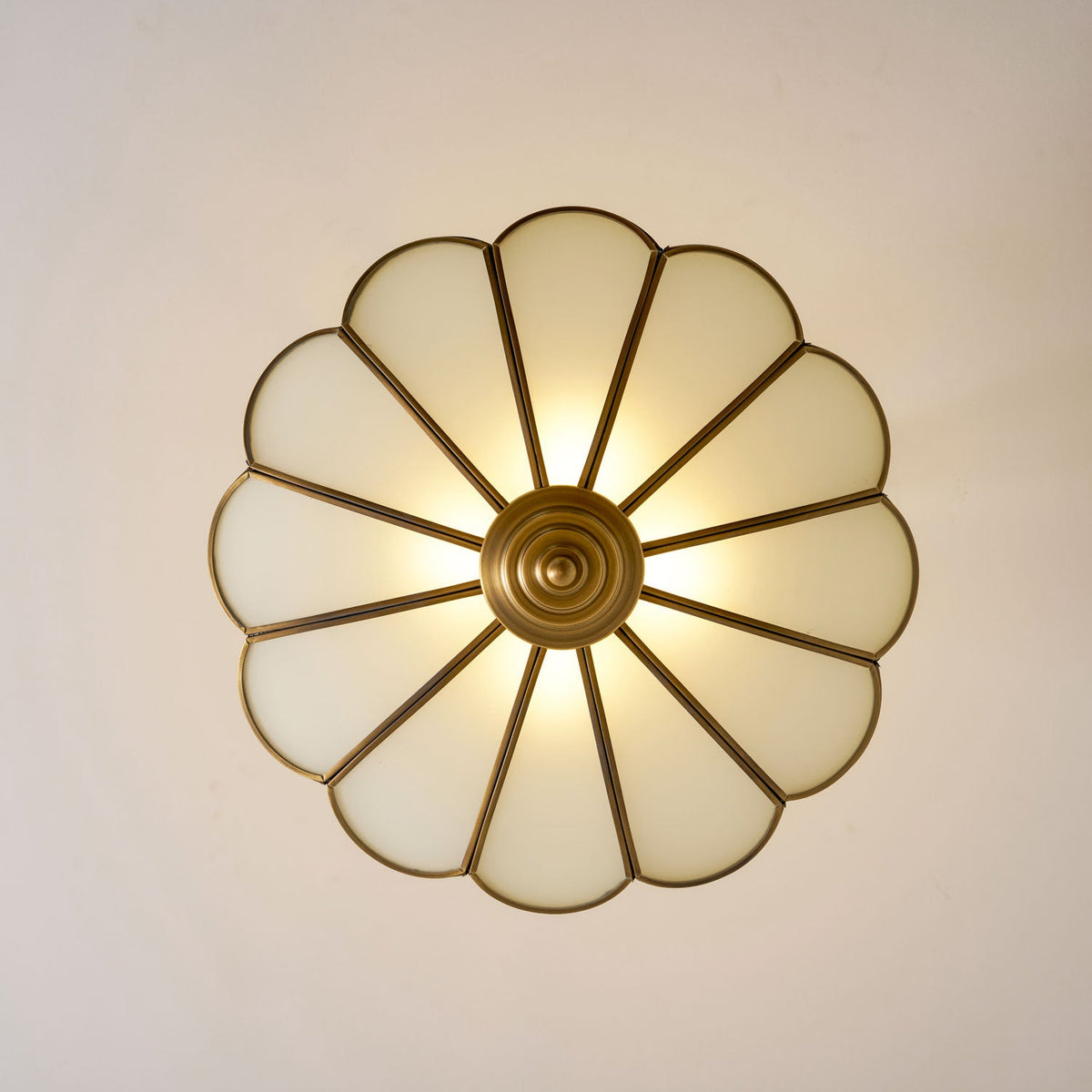 Seen from directly below, the scalloped frosted-glass petals of this Clérachic ceiling light glow warm around its brass centre.