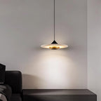 Clérachic flat disc pendant light glows above a dark leather bench in the corner of a plain grey room.