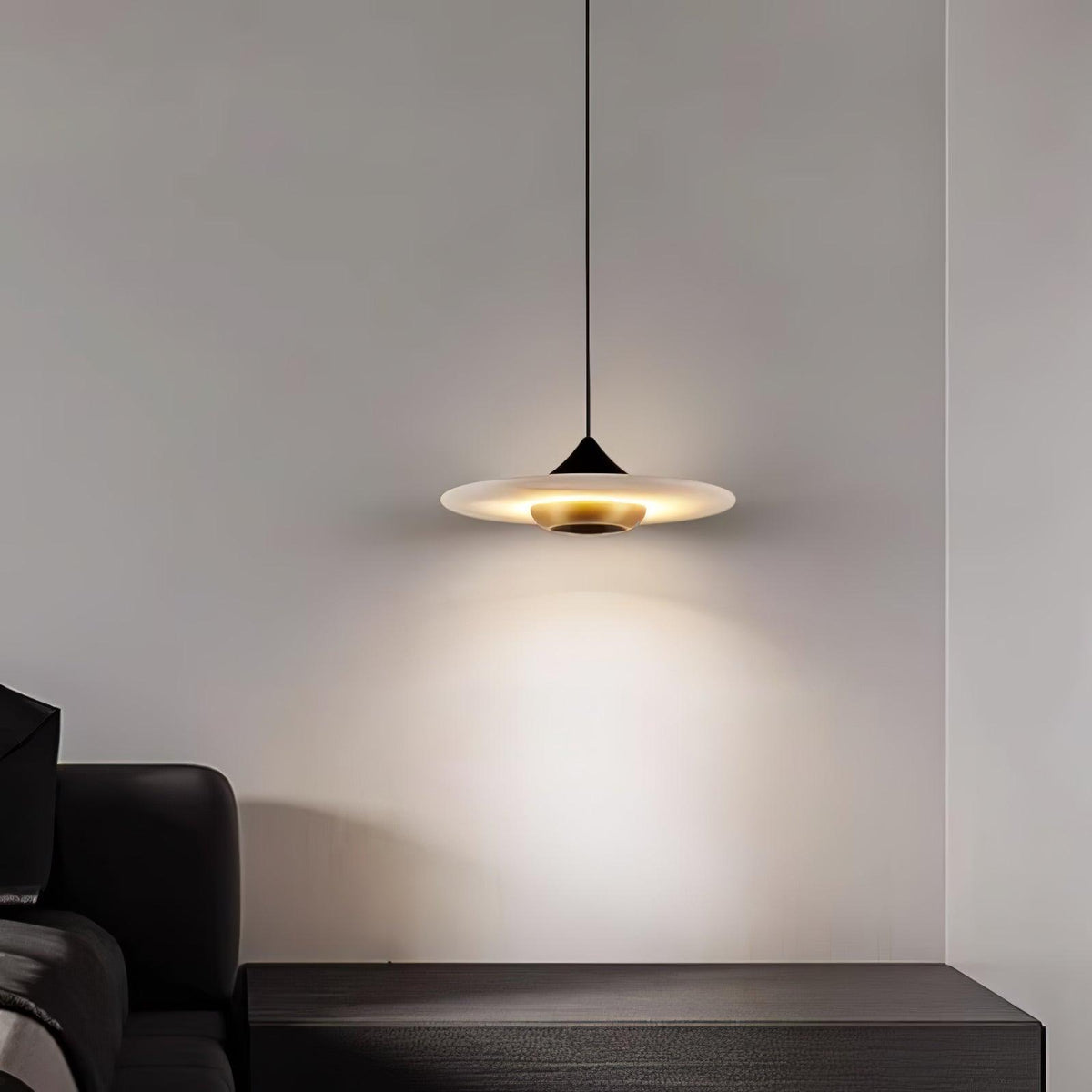 Clérachic flat disc pendant light glows above a dark leather bench in the corner of a plain grey room.