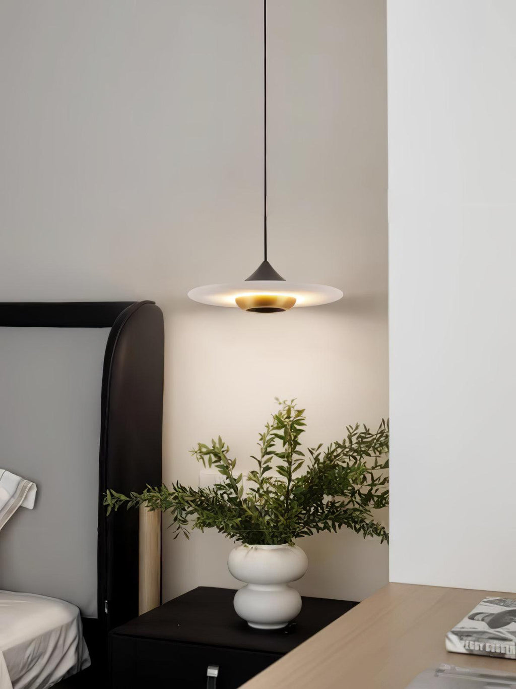 Clérachic white disc pendant glows above a dark nightstand beside a bed, with a white vase of greenery nearby.