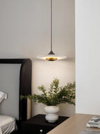 Clérachic white disc pendant glows above a dark nightstand beside a bed, with a white vase of greenery nearby.