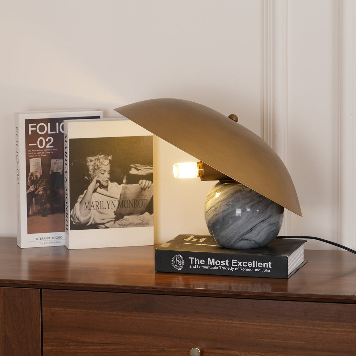 Clérachic lamp lit beside framed book covers on a walnut sideboard, its tilted shade angled toward the light.