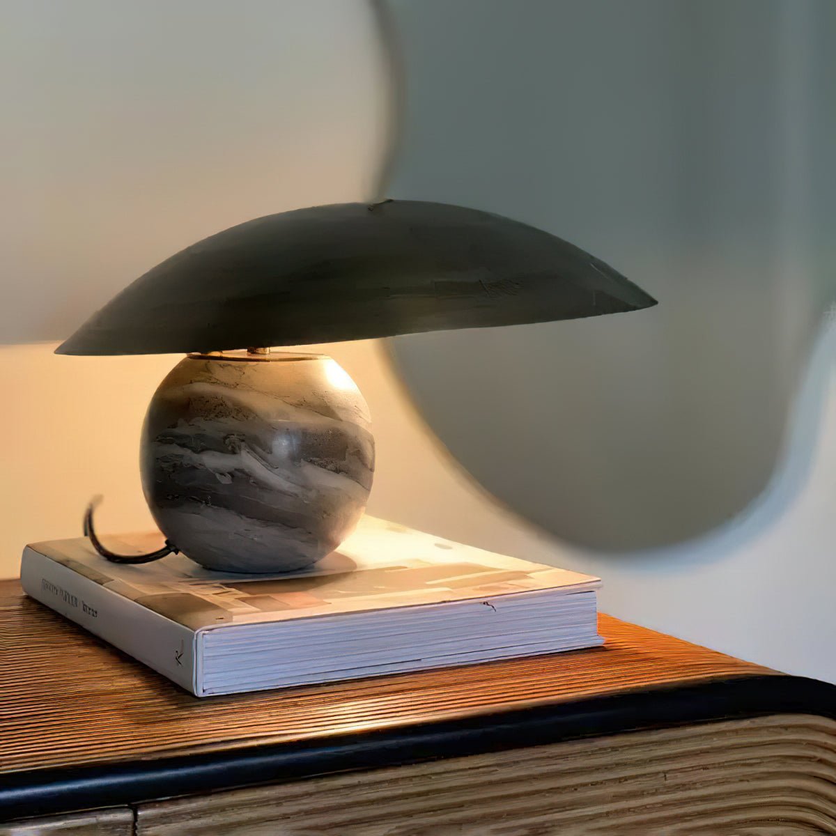 Clérachic table lamp with a tilted bronze dome shade on a marbled sphere base, resting on a book on a woven console.
