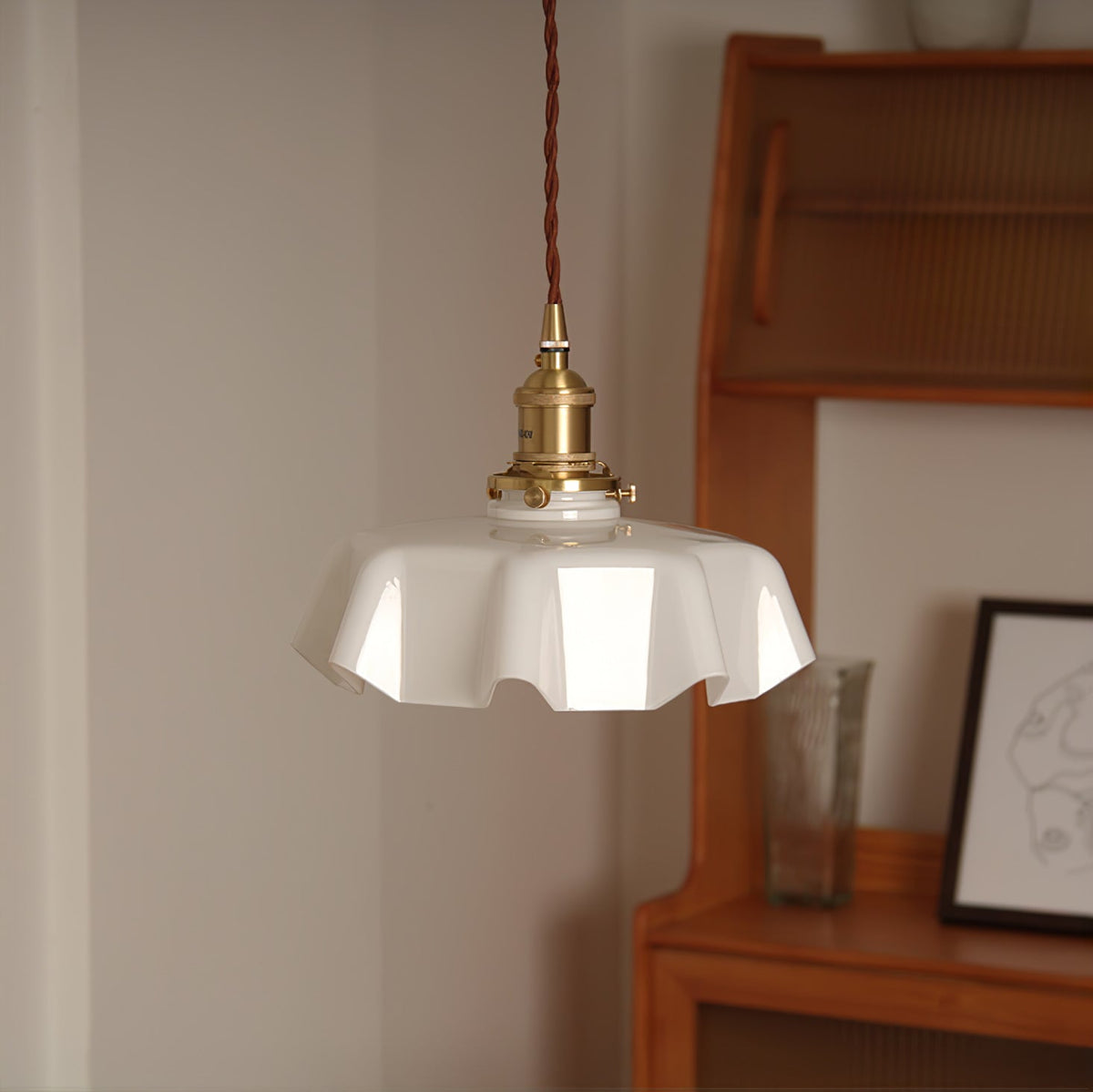 Beside a wooden cabinet and small framed print, this Clérachic pendant's white pleated glass shade hangs from its brass fitter.