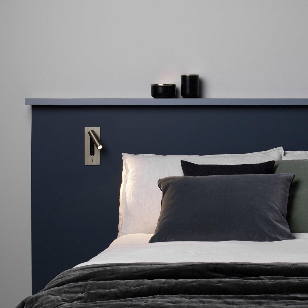Above a navy headboard with small candles on a shelf, this Clérachic reading light in nickel sits mounted on the wall.