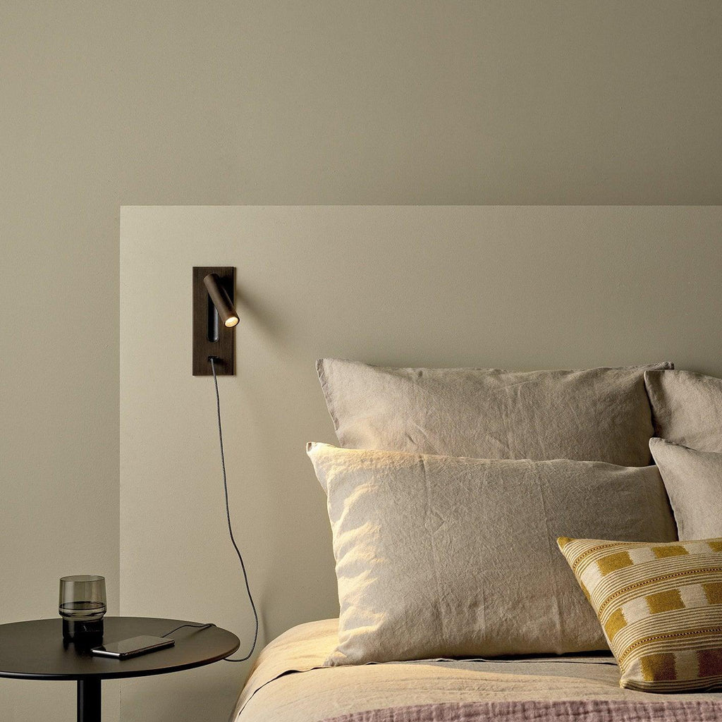 Beside a bed with linen cushions and a round side table, this Clérachic reading light in bronze sits unlit on an olive wall.