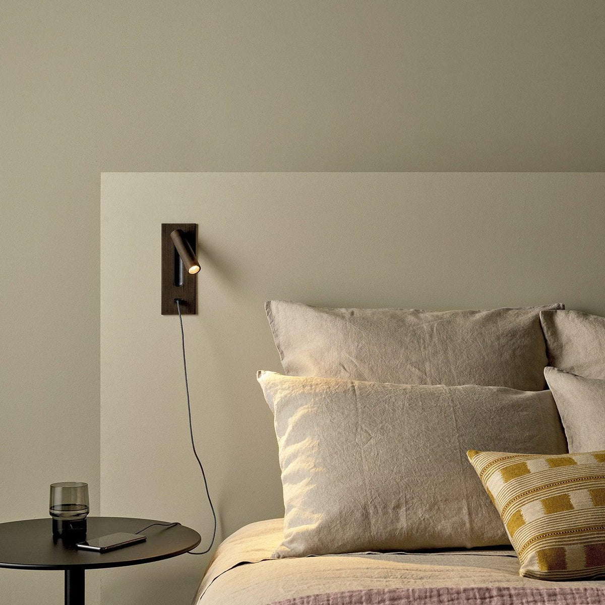 Beside a bed with linen cushions and a round side table, this Clérachic reading light in bronze sits unlit on an olive wall.
