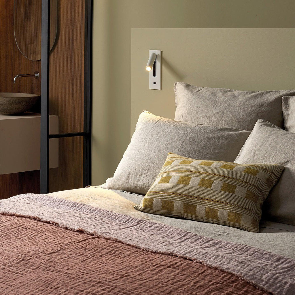 Mounted above a bed with olive and cream cushions, this Clérachic reading light in nickel sits on a warm-toned wall.