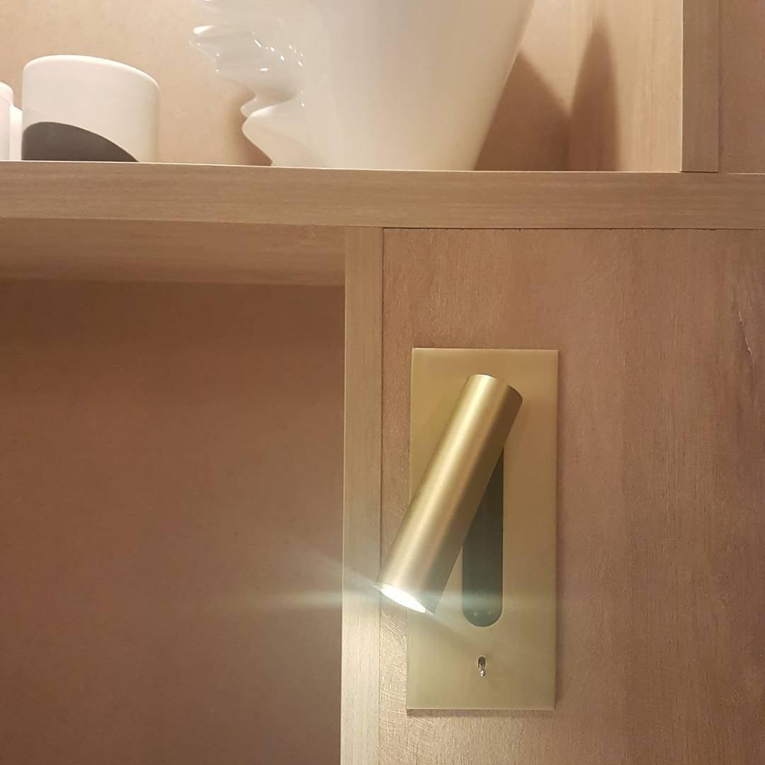 Lit and angled downward, this Clérachic reading light in copper is mounted in a wood-panelled bedroom nook above a shelf.
