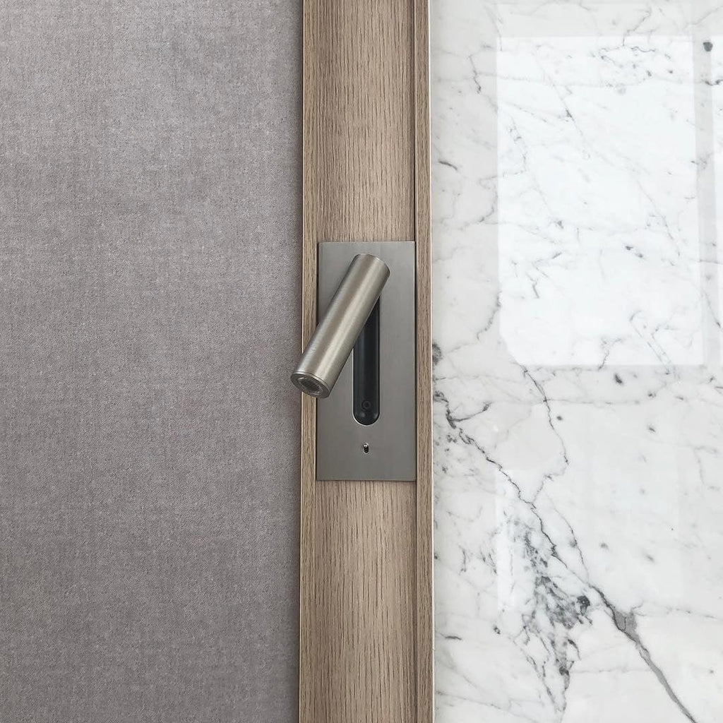Mounted on a grey fabric headboard beside a marble wall, this Clérachic reading light in nickel sits unlit.