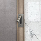 Mounted on a grey fabric headboard beside a marble wall, this Clérachic reading light in nickel sits unlit.