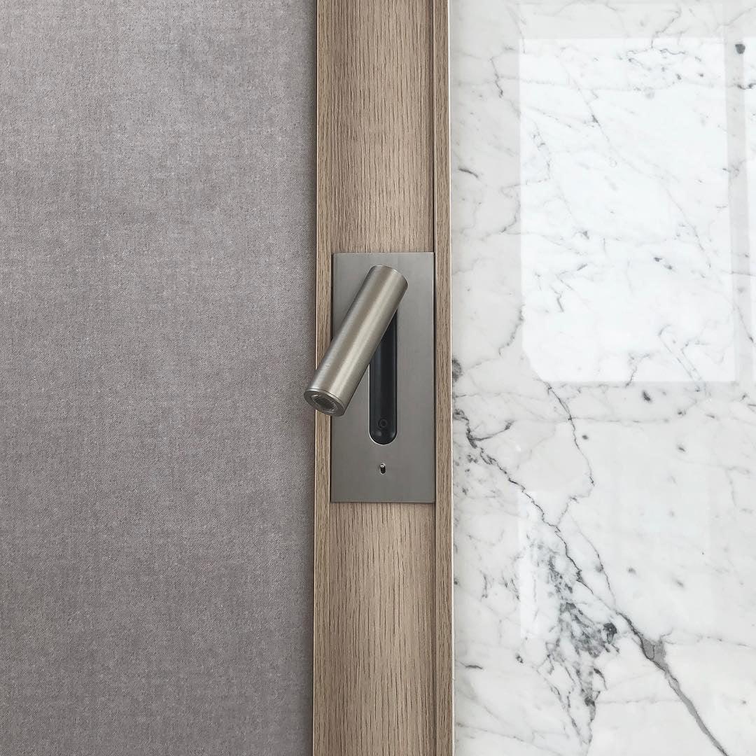 Mounted on a grey fabric headboard beside a marble wall, this Clérachic reading light in nickel sits unlit.