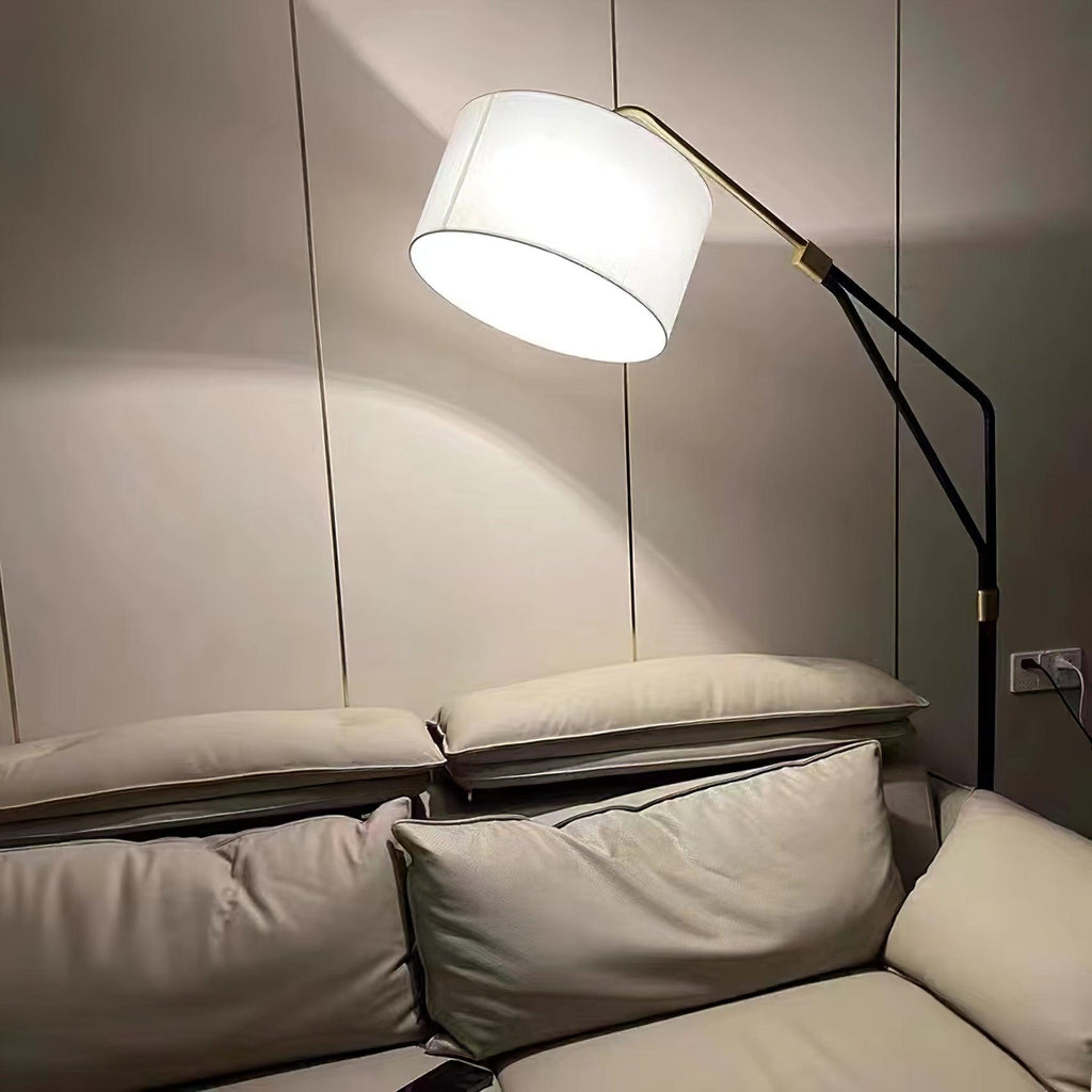 Clérachic black arc floor lamp glowing warm in a dim, dark-walled room, arched low over a taupe leather sofa by a socket.