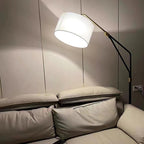 Clérachic black arc floor lamp glowing warm in a dim, dark-walled room, arched low over a taupe leather sofa by a socket.