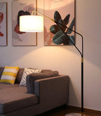 Clérachic black arc floor lamp arched over a taupe sofa with yellow patterned cushions, beside framed botanical prints.