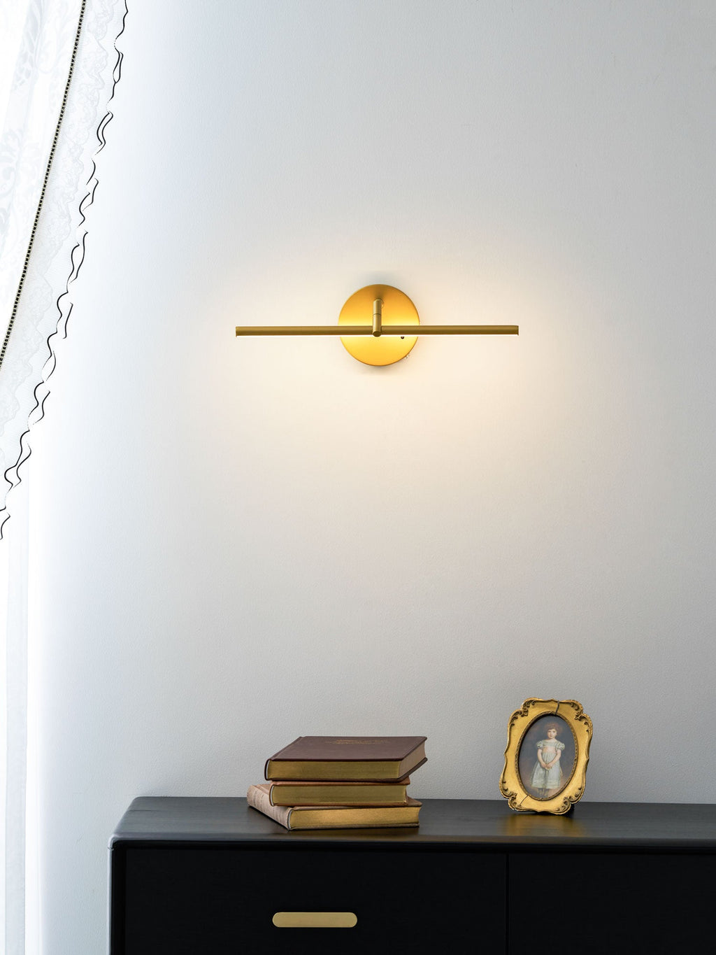 This gold-finish Clérachic wall light glows above a black dresser holding stacked books and a small oval gilt frame.