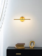 This gold-finish Clérachic wall light glows above a black dresser holding stacked books and a small oval gilt frame.