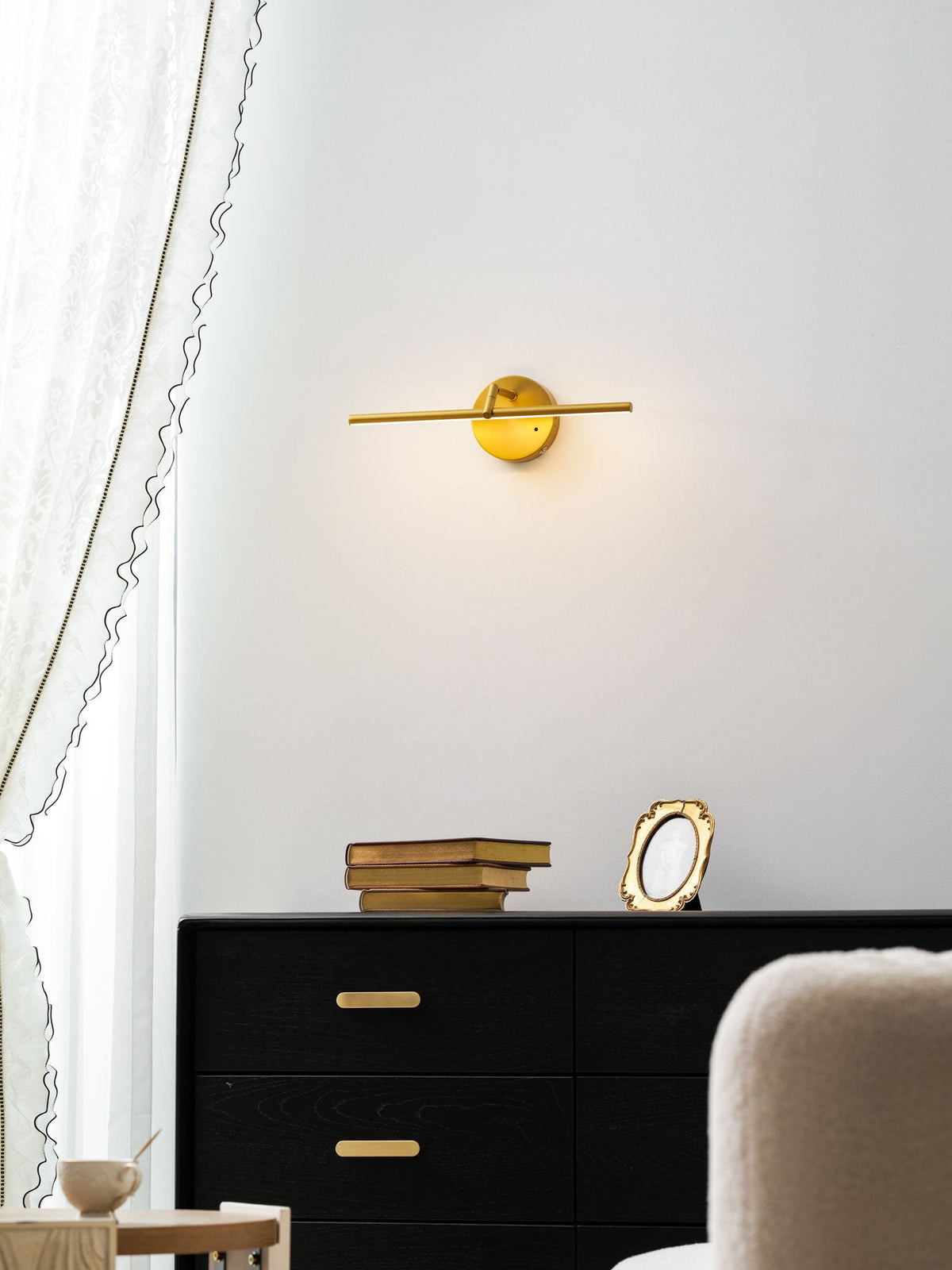 At a low angle, this gold-finish Clérachic wall light glows warmly above a black dresser beside a cream armchair.