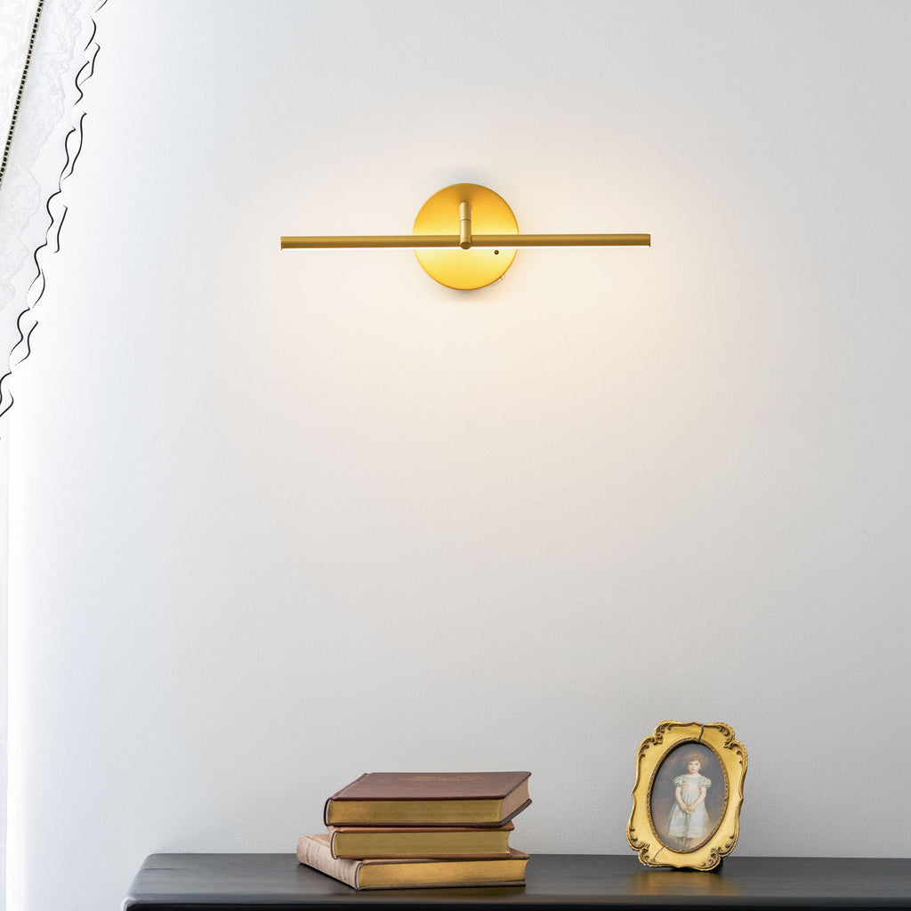 Lit gold-finish wall light from Clérachic above a black dresser with books and a framed portrait, a sheer curtain draped beside it.