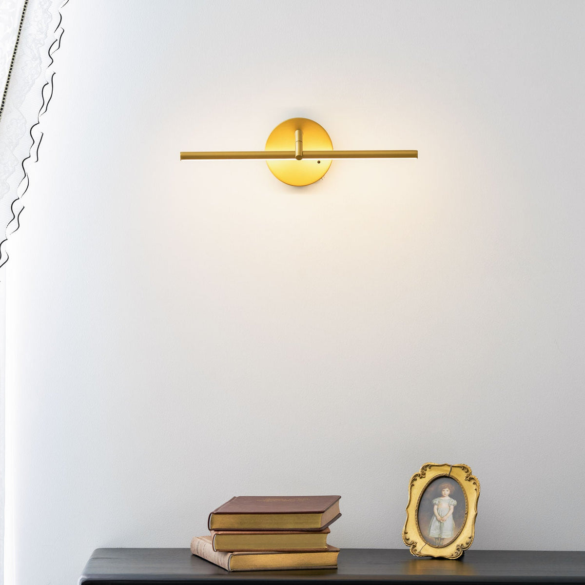 Lit gold-finish wall light from Clérachic above a black dresser with books and a framed portrait, a sheer curtain draped beside it.