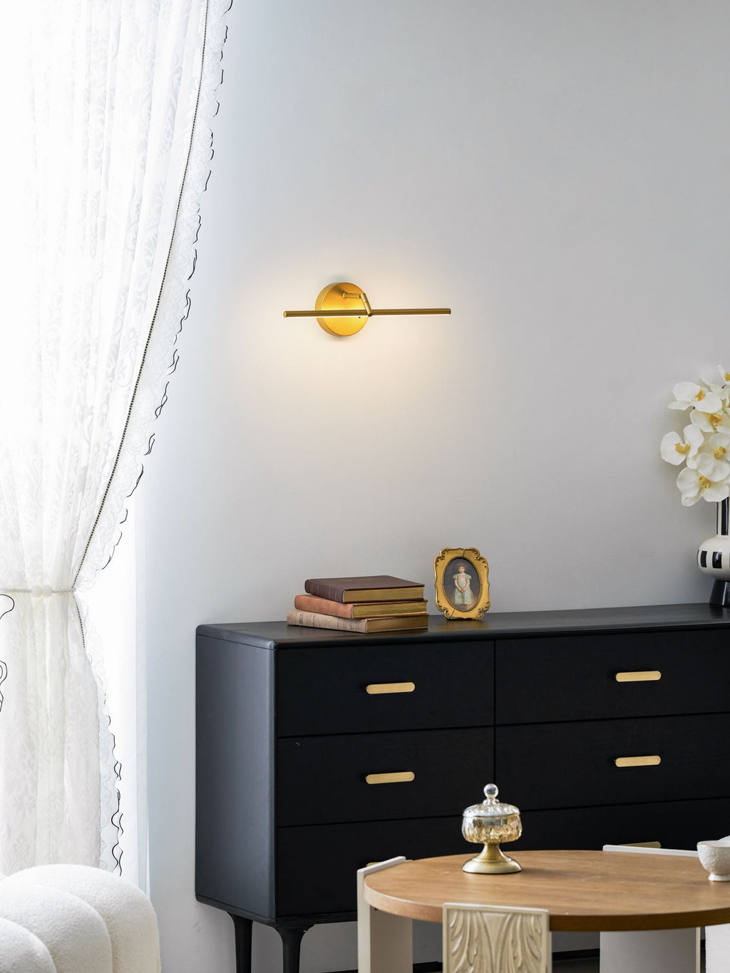 This gold-finish Clérachic wall light glows above a black dresser and a round table set with a cloche and an orchid, curtain beside it.