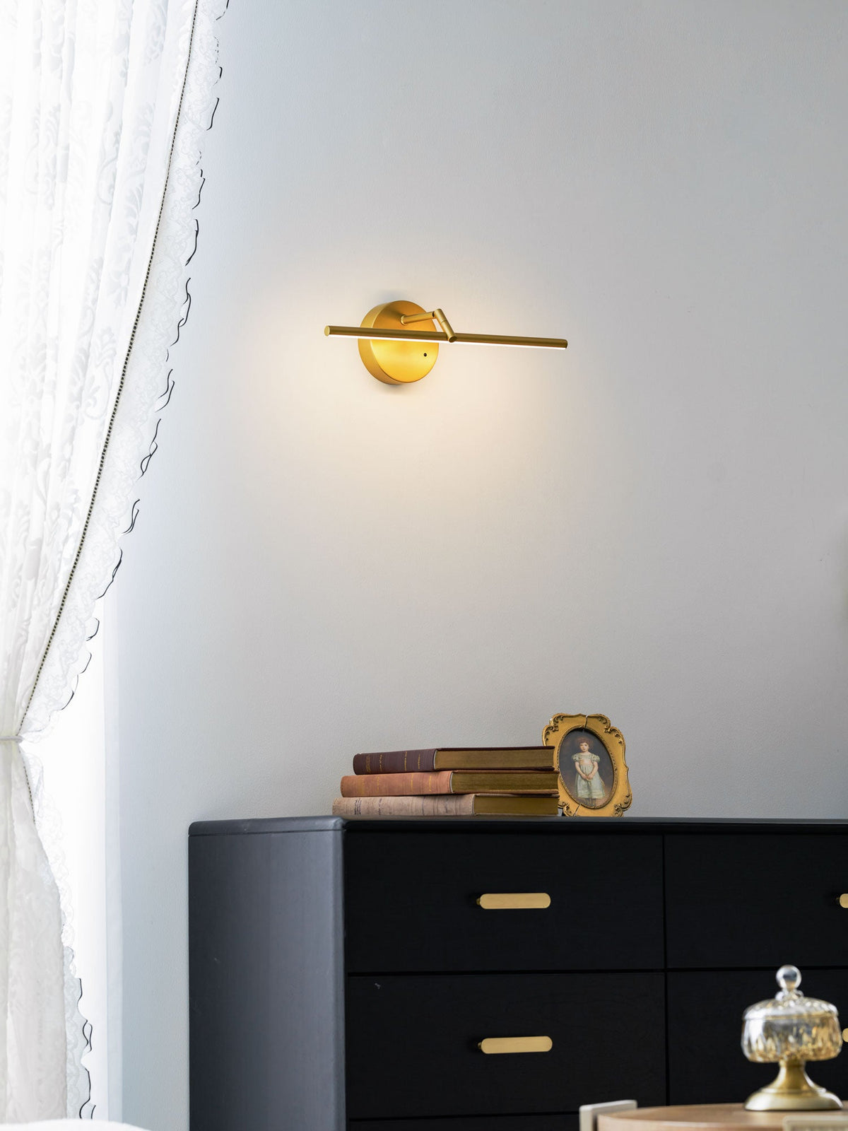 Mounted above a black dresser with brass drawer pulls, this gold-finish Clérachic wall light glows beside stacked books and a framed portrait.