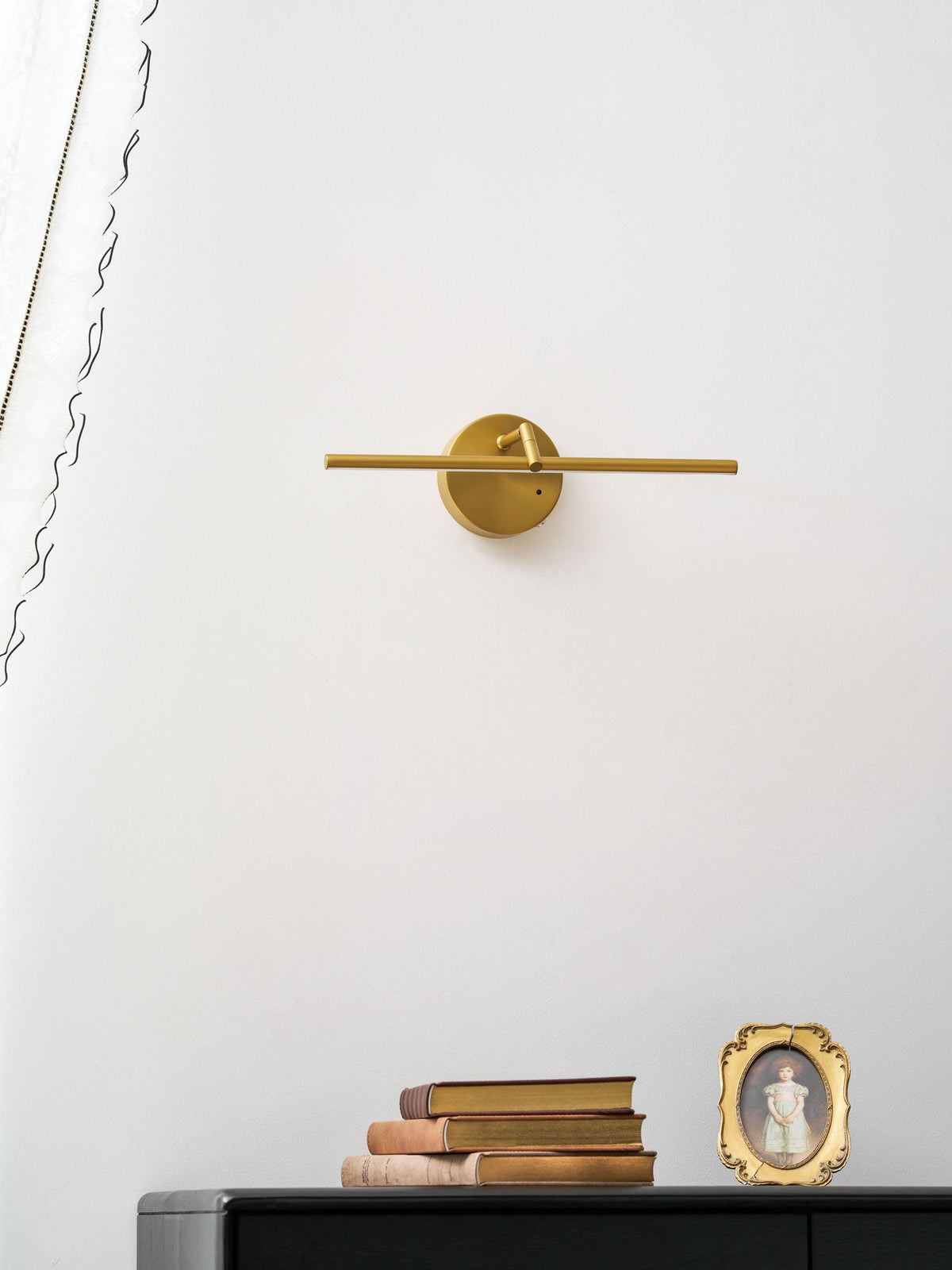Unlit gold-finish wall light from Clérachic above a black dresser with stacked books and a small framed portrait, curtain at the edge.