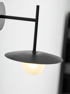 A low, close angle on this black Clérachic wall light, its disc shade glowing warmly.