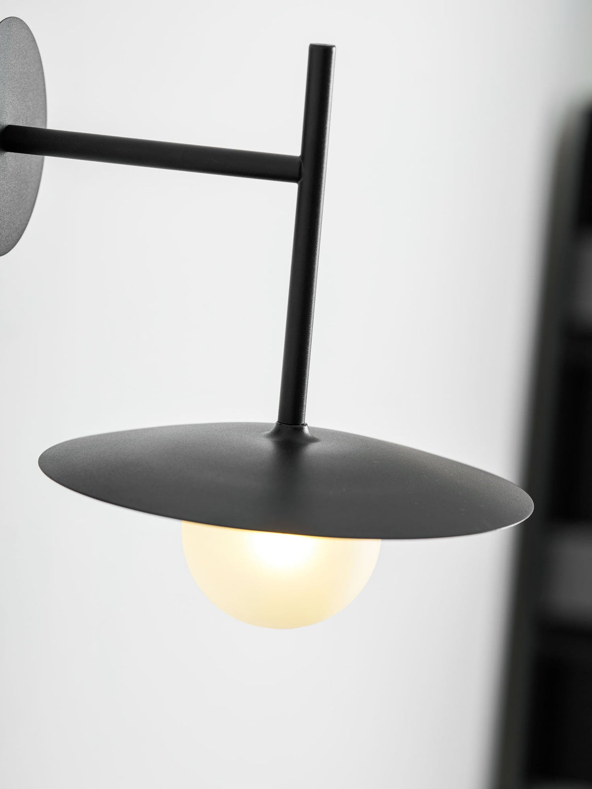 A low, close angle on this black Clérachic wall light, its disc shade glowing warmly.