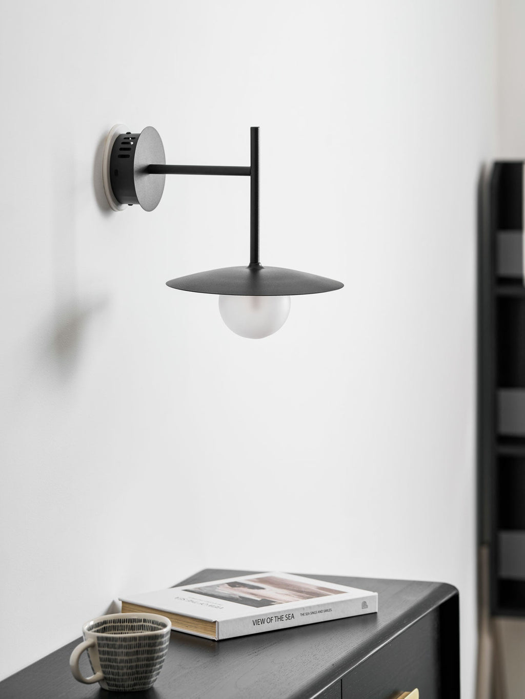 Grey Clérachic wall light shown unlit above a dark dresser set with a cup and a book.
