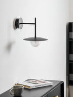 Grey Clérachic wall light shown unlit above a dark dresser set with a cup and a book.