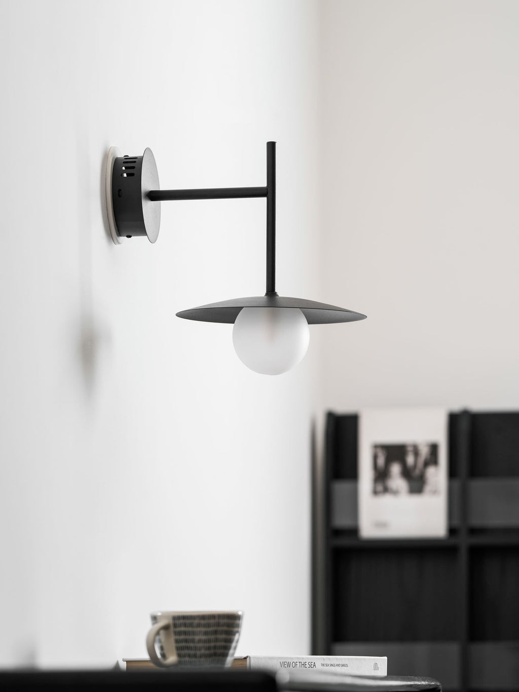 White Clérachic wall light shown unlit above a dark dresser holding a magazine and a cup.