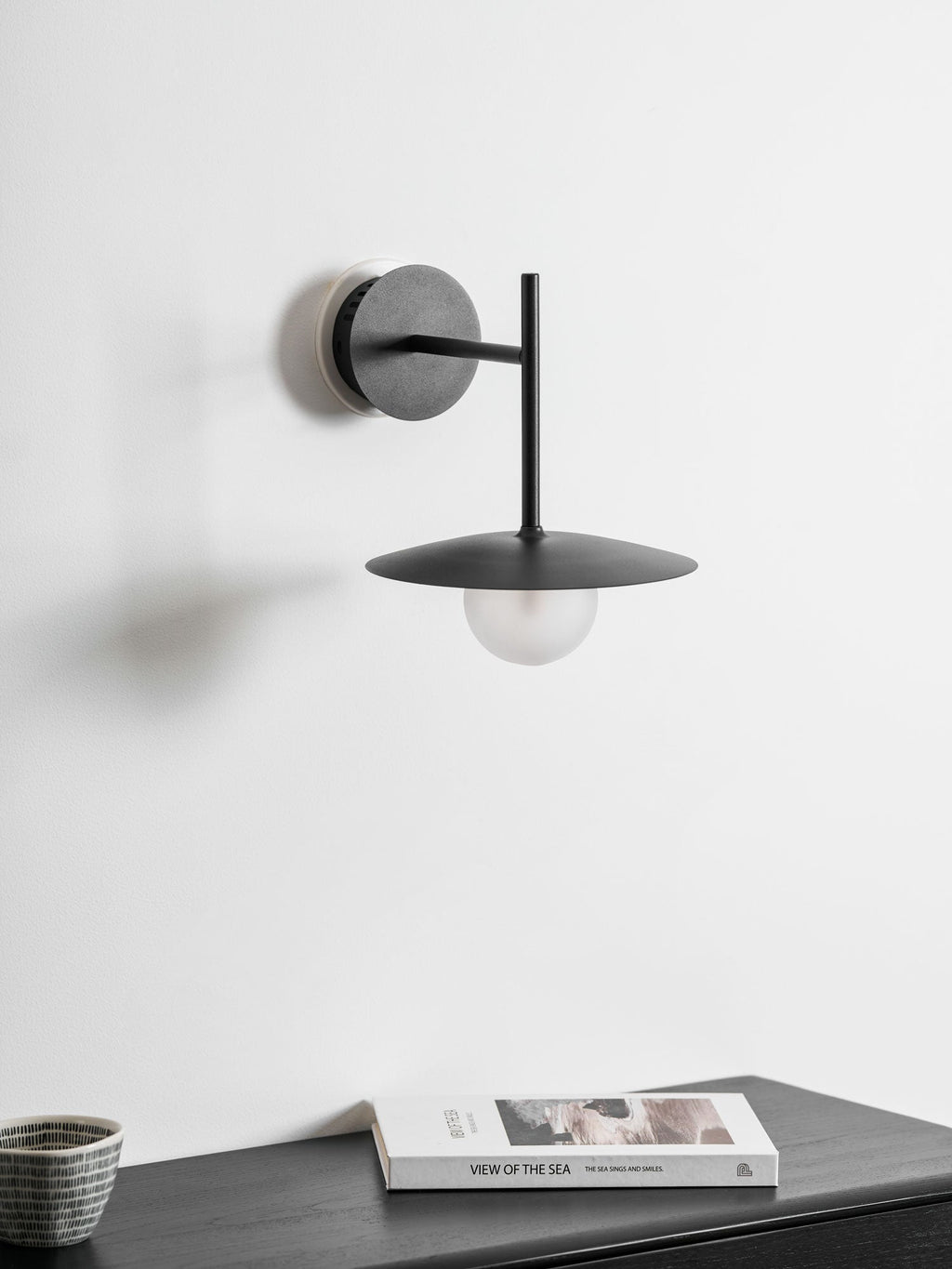 Grey Clérachic wall light shown unlit above a dark dresser stacked with a magazine.