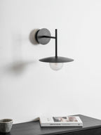 Grey Clérachic wall light shown unlit above a dark dresser stacked with a magazine.
