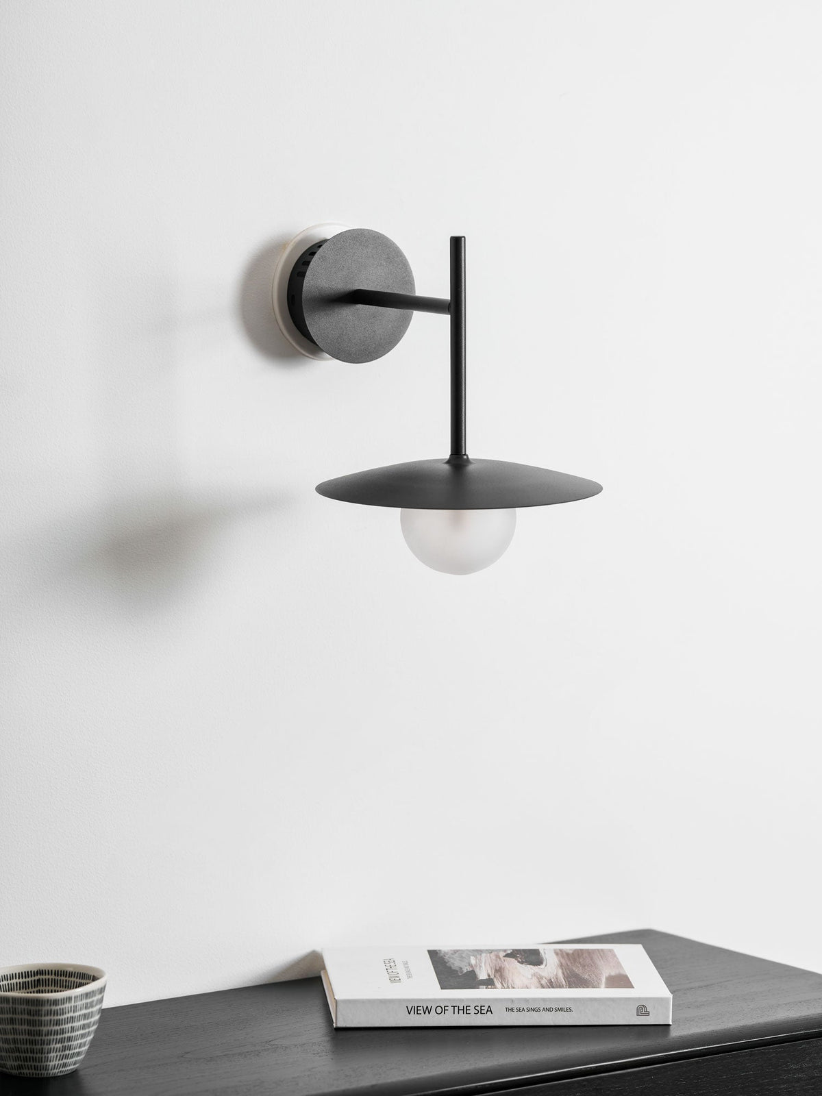 Grey Clérachic wall light shown unlit above a dark dresser stacked with a magazine.