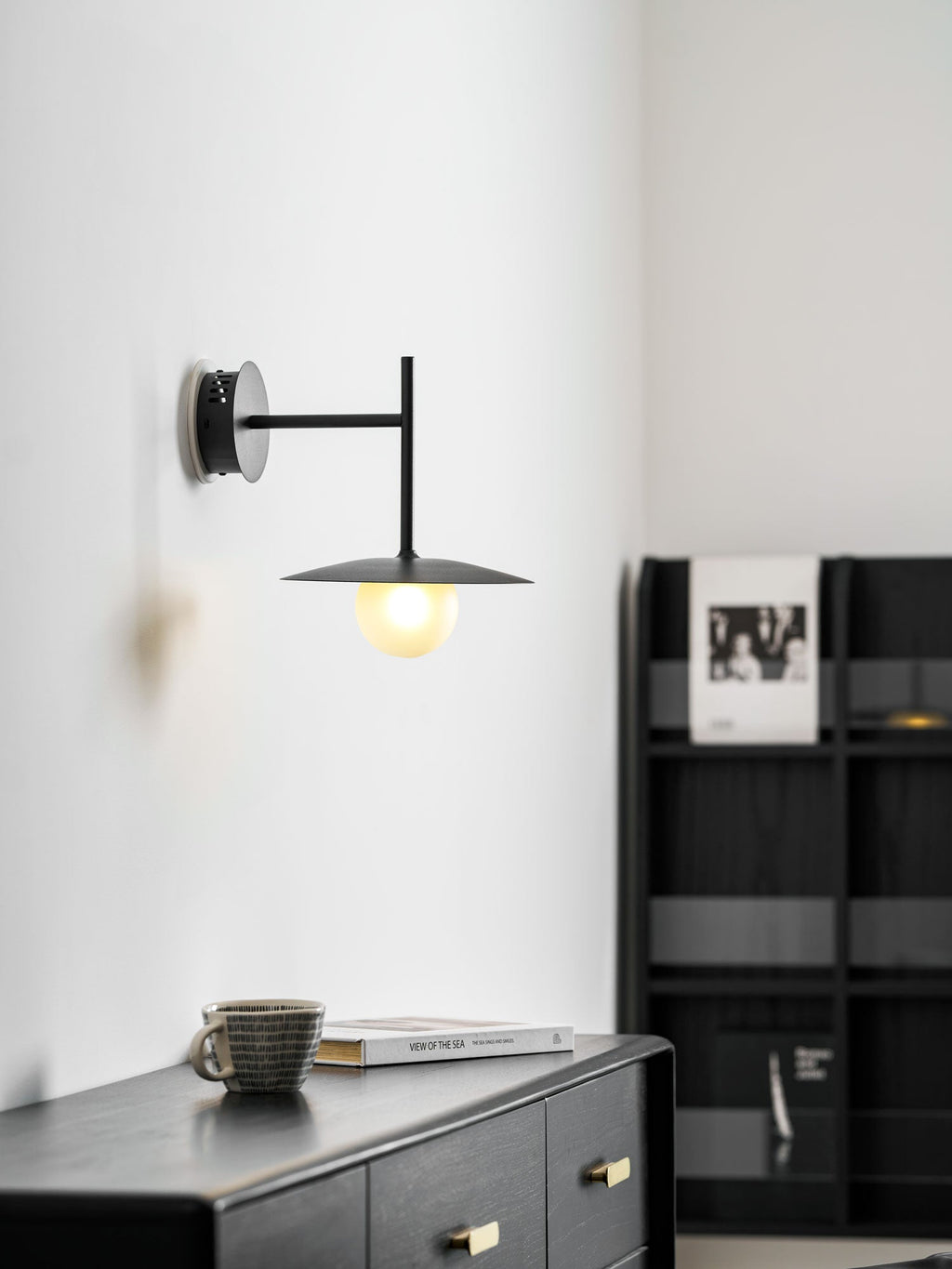 White Clérachic wall light glows above a dresser beside a black bookshelf lined with books.