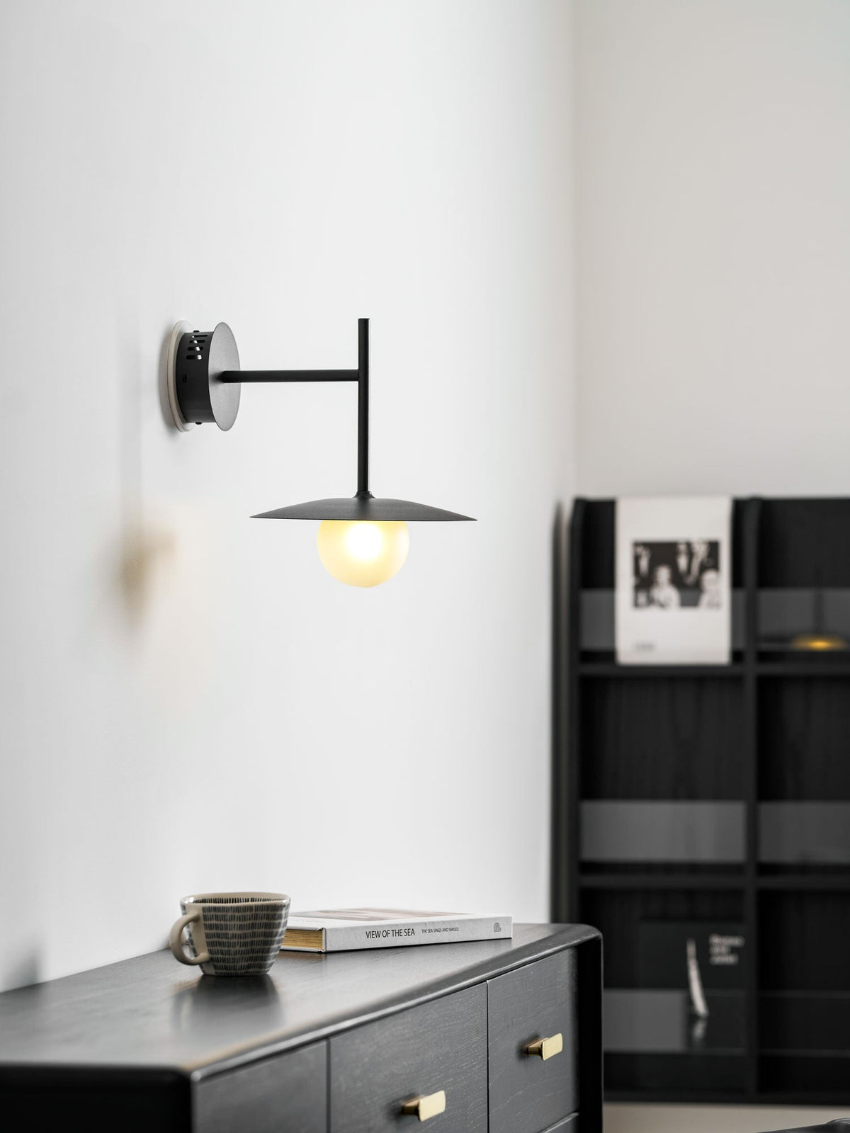 White Clérachic wall light glows above a dresser beside a black bookshelf lined with books.