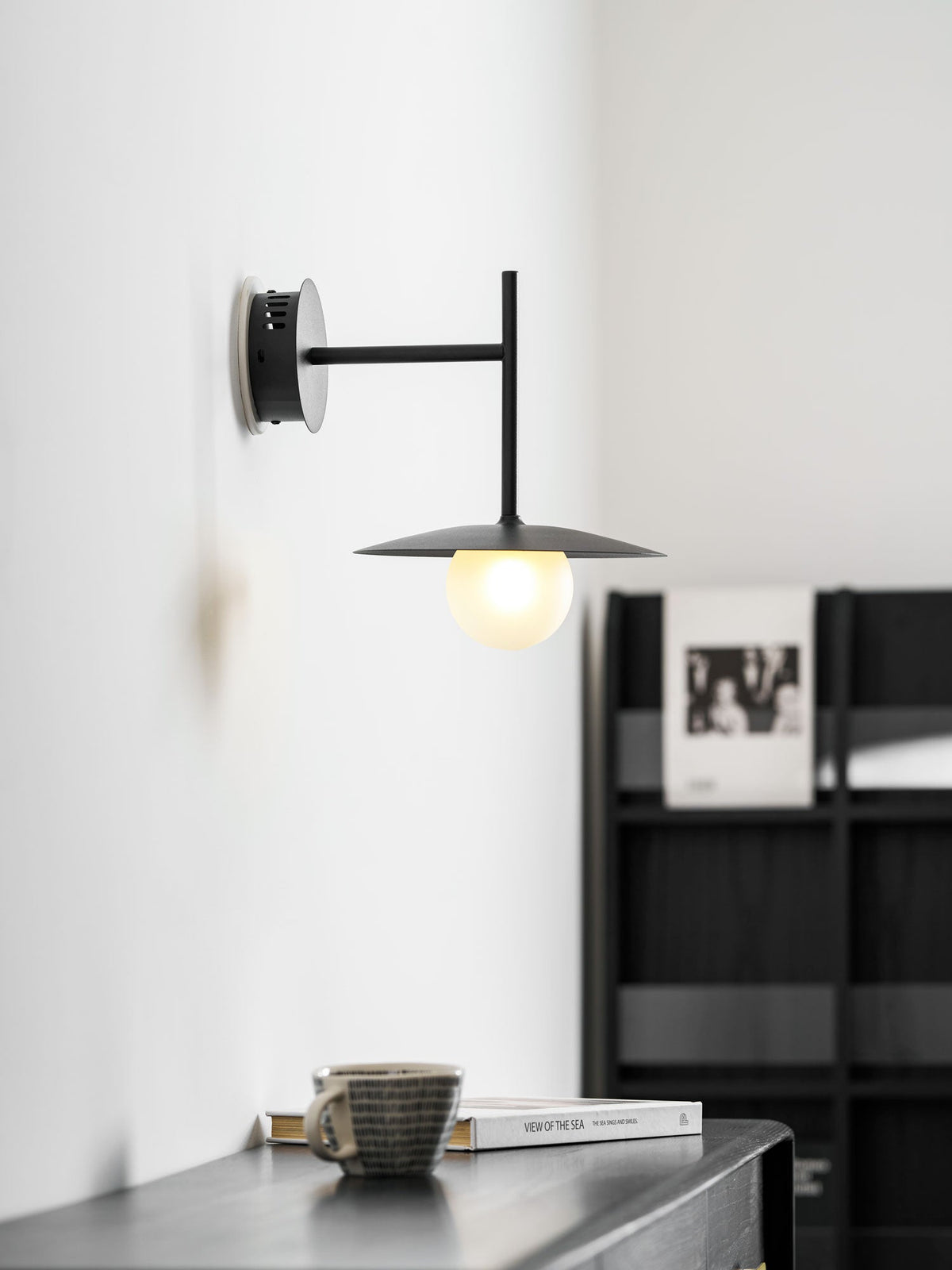 Grey Clérachic wall light glows above a dresser holding a cup and a stacked book.