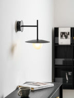 Black Clérachic wall light glows beside a black bookshelf, its disc shade lit warmly.