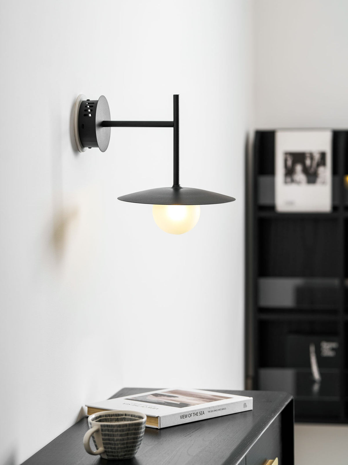 Black Clérachic wall light glows beside a black bookshelf, its disc shade lit warmly.