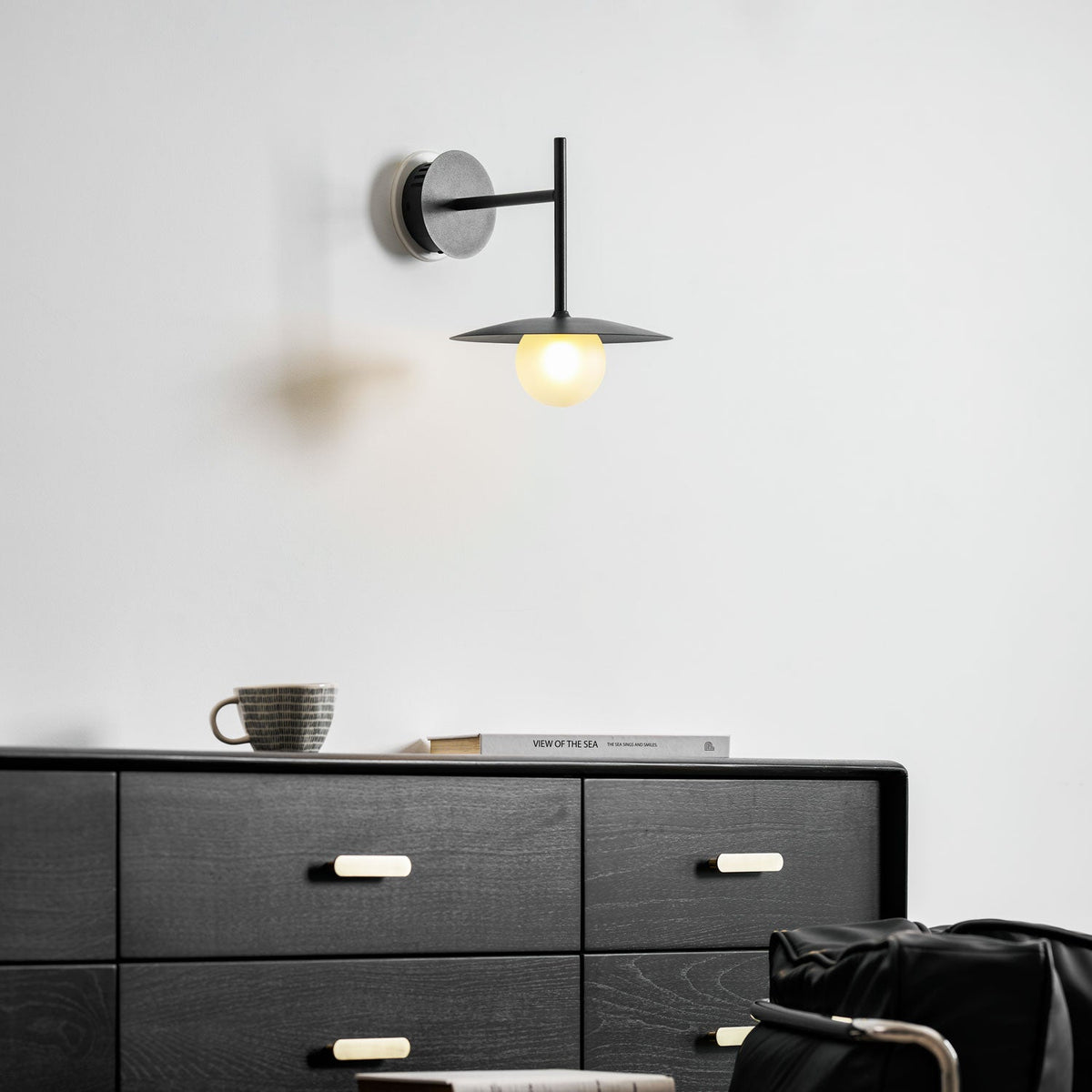 Grey Clérachic wall light glows above a dresser set with a mug and folded books.