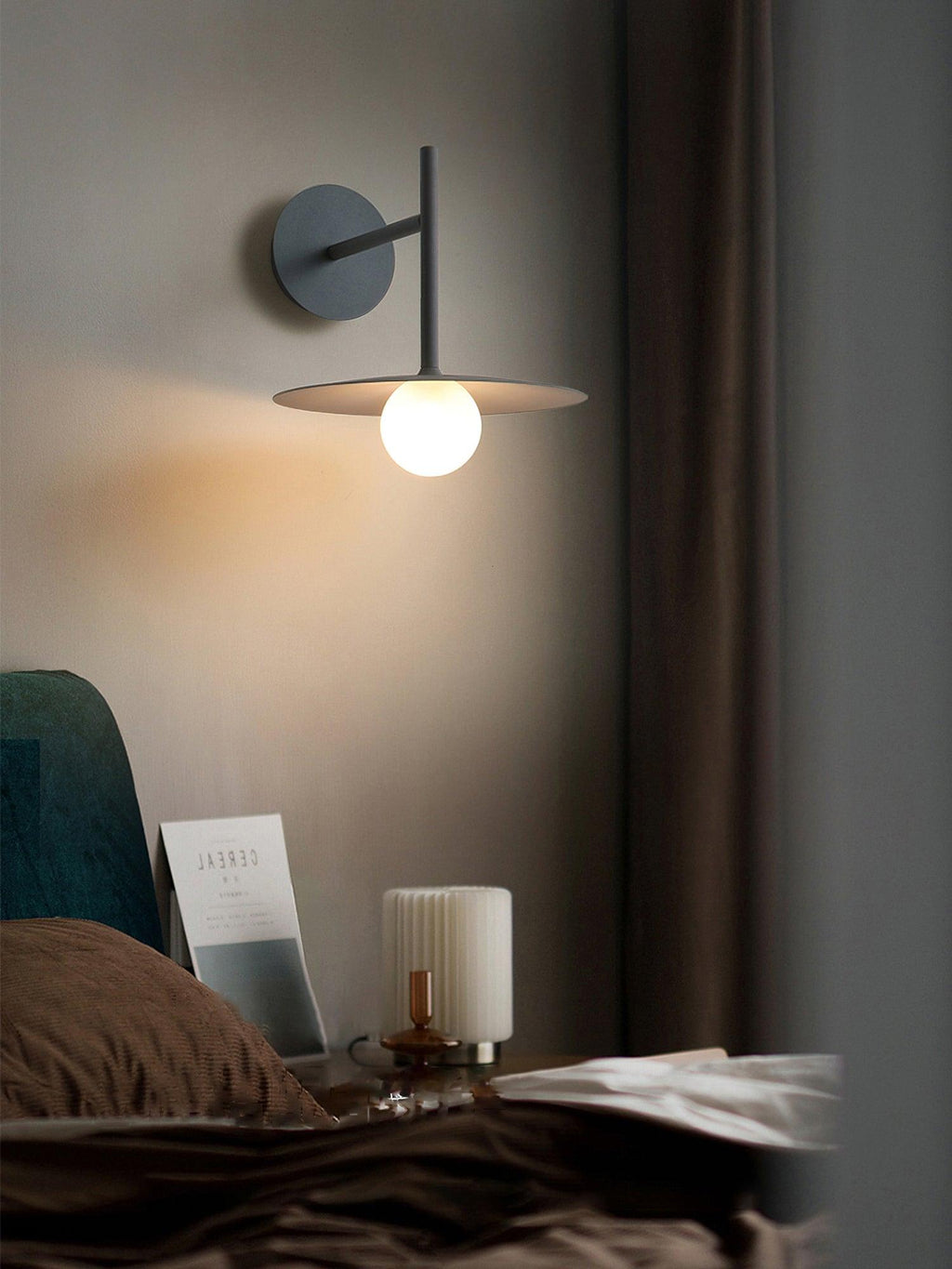 This black Clérachic wall light glows beside a dark curtain, a book resting on the nightstand below.