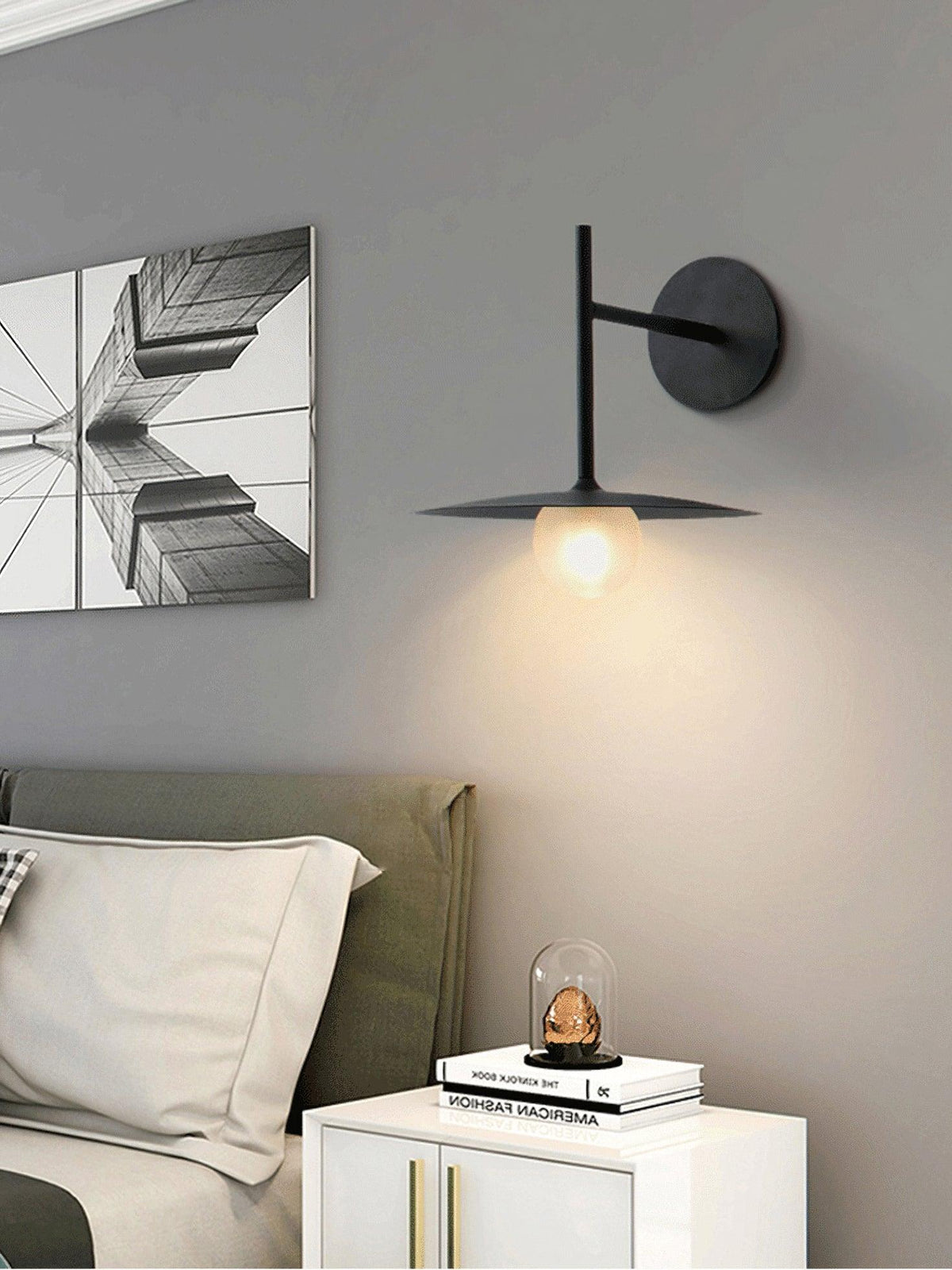 Above a white nightstand holding books and a glass dome, this black Clérachic wall light glows.