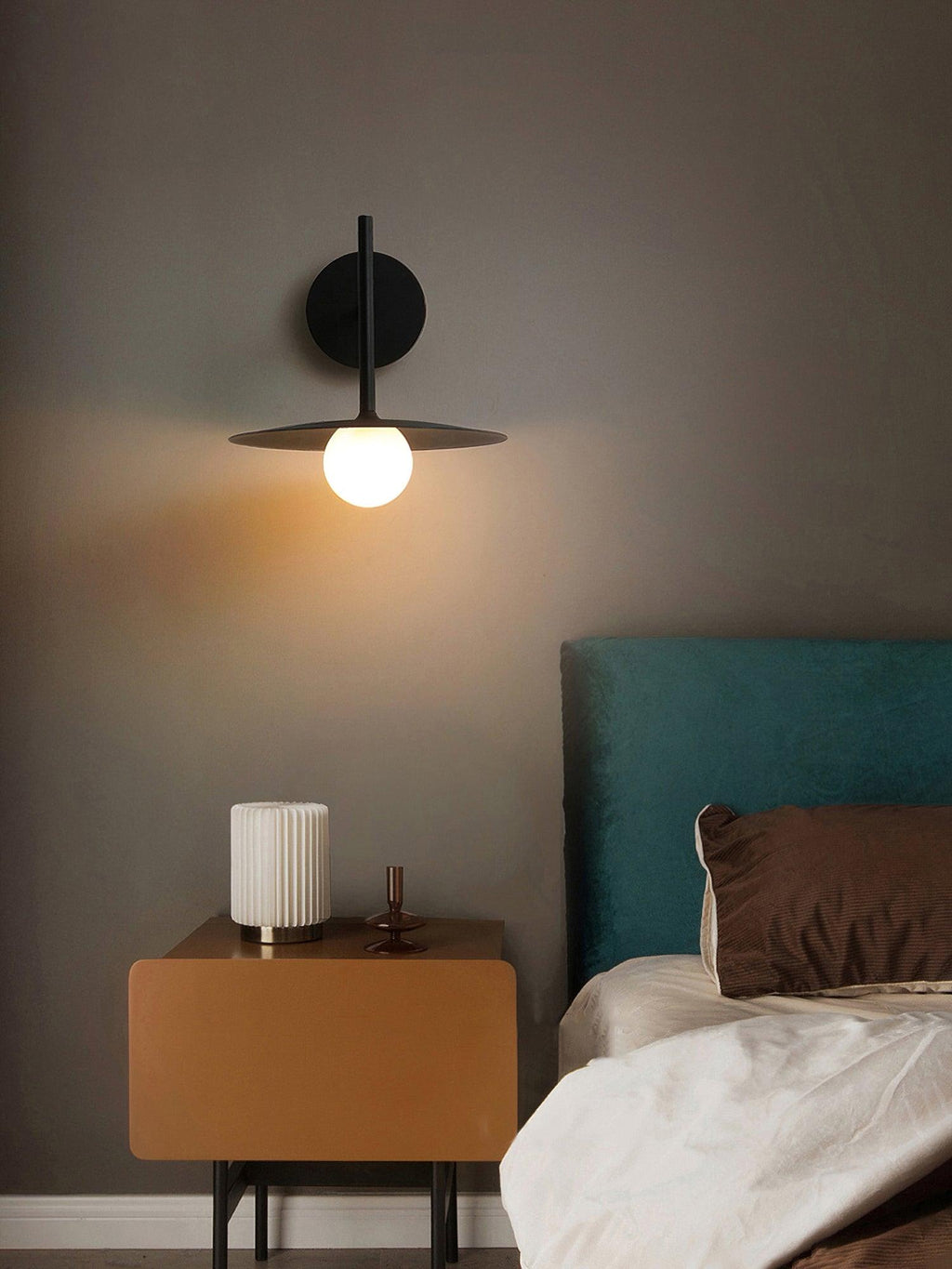 Above a tan nightstand, this black Clérachic wall light glows beside a teal upholstered headboard.