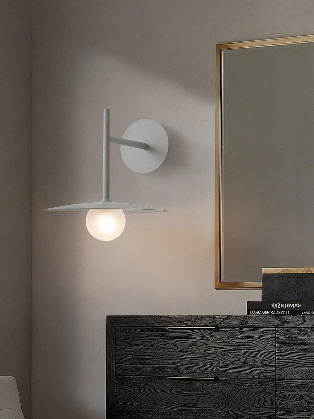 Beside a gold-framed mirror, this white Clérachic wall light glows above a dark wood dresser.