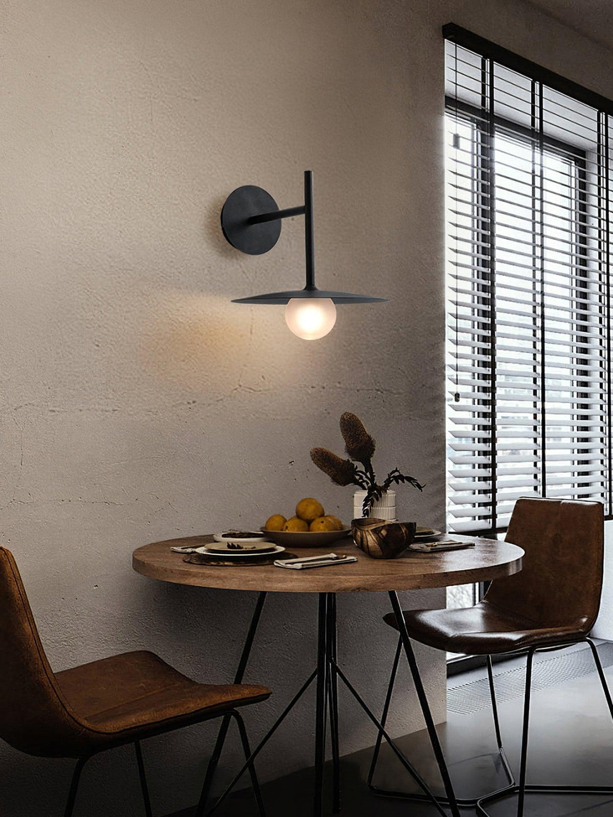 Black Clérachic wall light lit above a round dining table set with leather chairs.