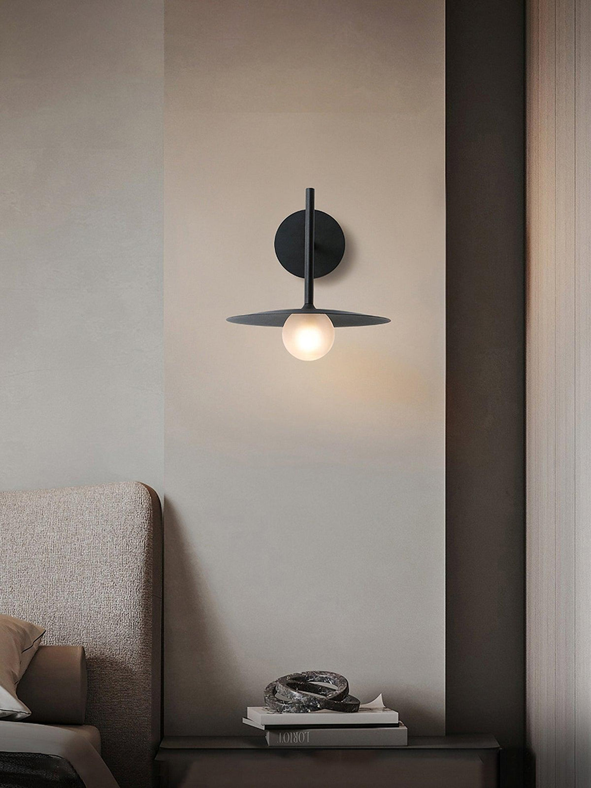 In a bedroom corner, this black Clérachic wall light glows beside a stack of books.